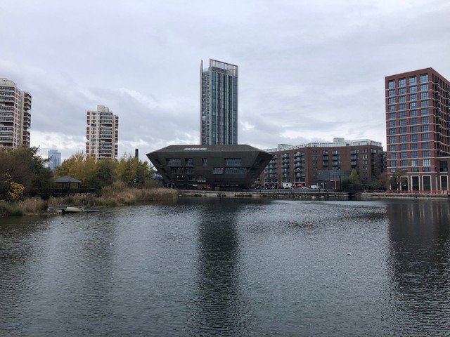 Canada library building in front of a lake. The building is narrower at the bottom and wider at the top, like a rhombus (or a star wars sand crawler.) Behind the building are square buildings and  skyscrapers.