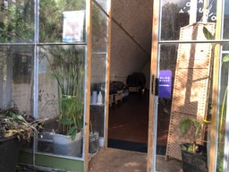 Brick arch with a large room underneath. Greenhouse stlye front has plants in pots. Blankets are on a ben in the mainly empty room. Sign on the glass door says y-oga studio.