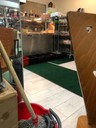A cafe counter, a mop and bucket are in the foreground. Peeping out from above a tall rack next to the counter, you can see the lime green headscarf of the Vegan Rasta