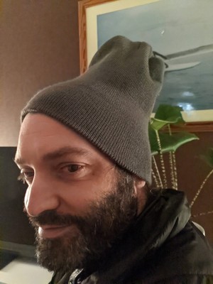 Bearded man in a grey beanie hat that clings to his fore head like a skull cap, but sticks out at the back like an idiot.