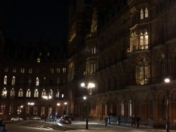 Night time view of a grand, old, brick and stone building for or five storeys high with ornate arched windows. A sweeping road concourse is in front. It looks like Hogwarts.
