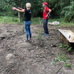 Two people stand in a clearing in a woodland. Some small trees have been planted with posts supporting them.