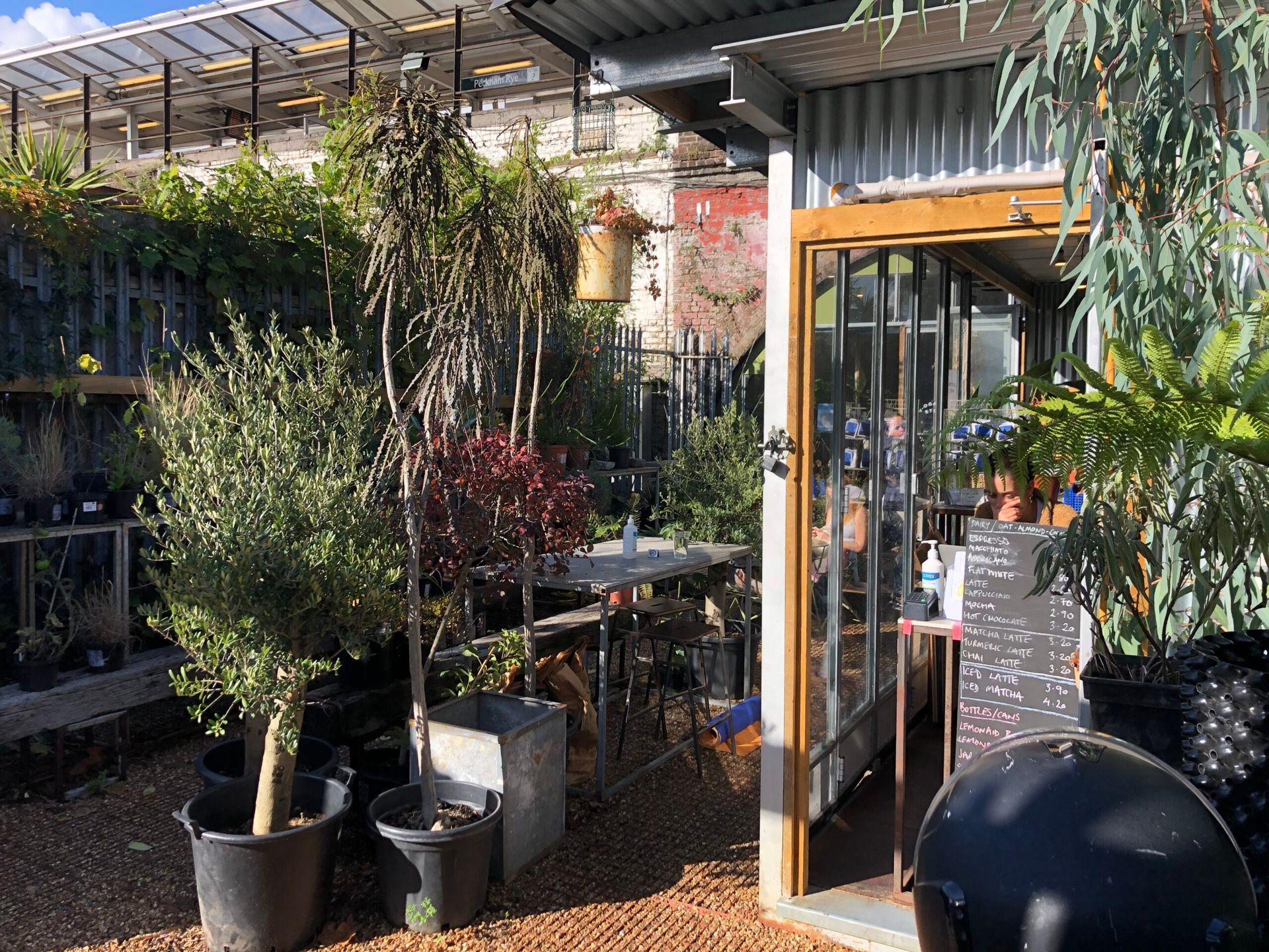 Plants in pots, but has a metal shack selling lattes etc, there are wooden tables. A grey wall in the background with a train station platform above. is actually a cafe