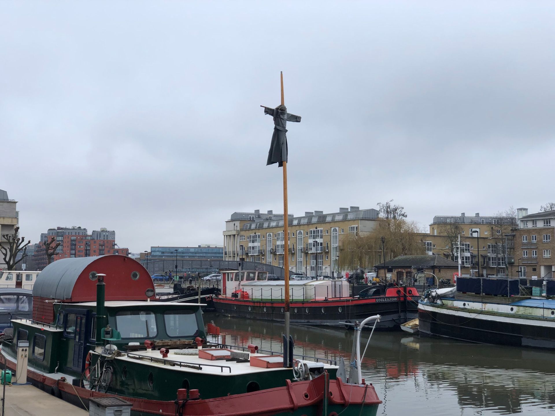 A scarecrow like figure hangs on a twenty foot high crusafix on the mast of an old green barge houseboat in a dock surrounded by other houseboats and modern blocks of flats.