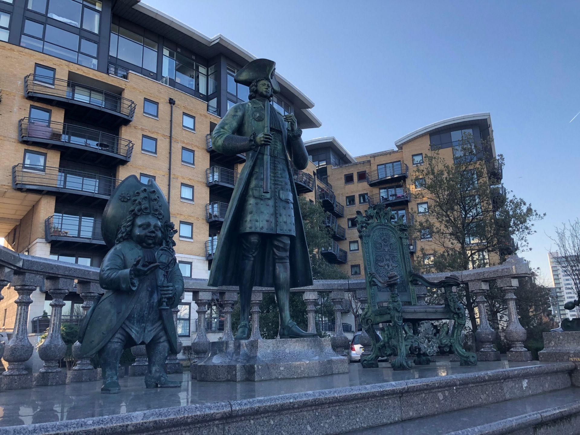 Statue of Peter The Great standing on a plinth. To the left is a statue of a court dwarf and to the right is a statue of a throne. All in Bronze.