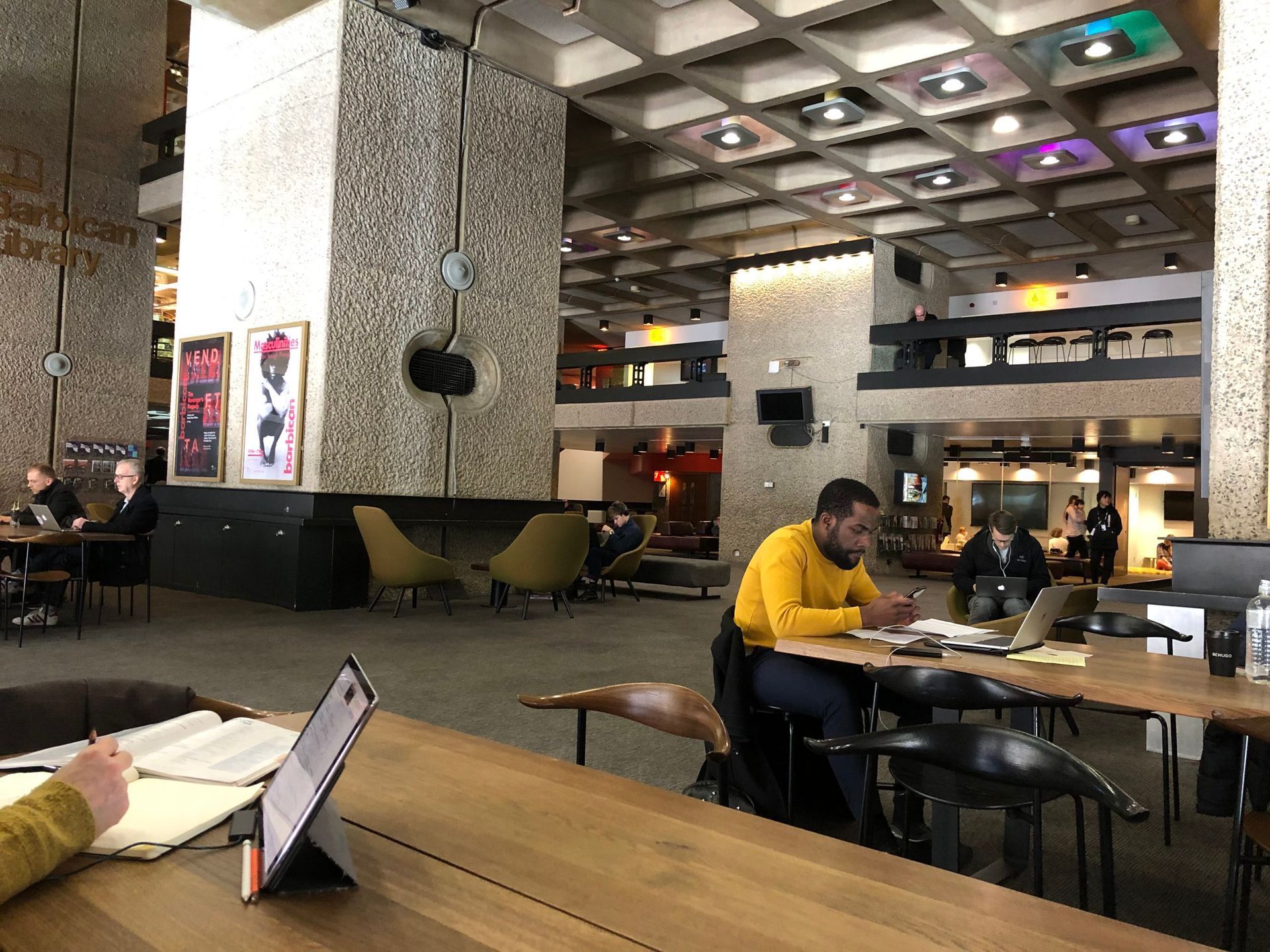 A large boxy granate hall, people work on laptops and drink coffee in the hall and on a mezzanine balcony above. Stylish furniture of different types. From long communal tables to arty armchairs to coffee counters stools.